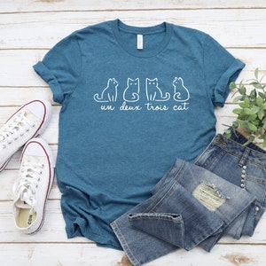 May include: A teal blue t-shirt with a white graphic of four cats and the text "un deux trois cat". The shirt is laid out on a white wooden surface with a pair of white sneakers and a pair of blue jeans.