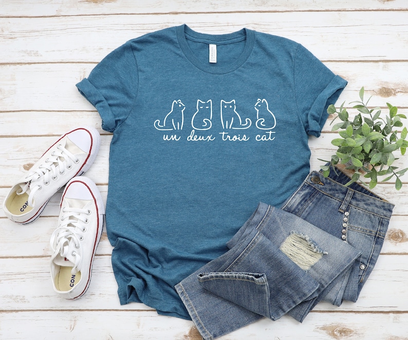 May include: A teal blue t-shirt with a white graphic of four cats and the text "un deux trois cat". The shirt is laid out on a white wooden surface with a pair of white sneakers and a pair of blue jeans.