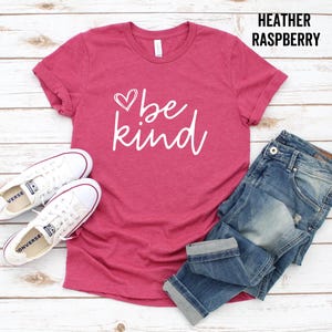 May include: A heather raspberry t-shirt with the text "be kind" in white lettering and a small heart graphic. The shirt is paired with blue jeans and white sneakers.