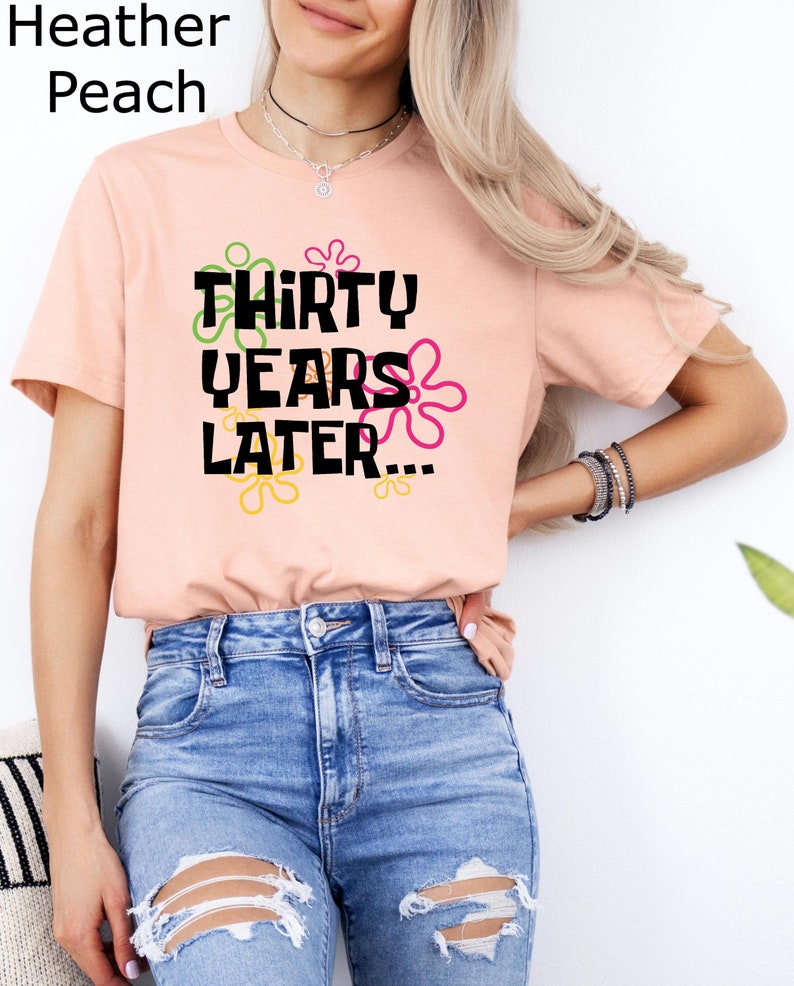 May include: A pink t-shirt with the text "THIRTY YEARS LATER..." in black lettering. The text is surrounded by colorful cartoon flowers.