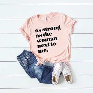 May include: A pink t-shirt with the text "as strong as the woman next to me." in black bold letters.