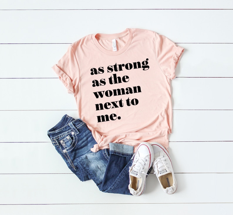 May include: A pink t-shirt with the text "as strong as the woman next to me." in black bold letters.