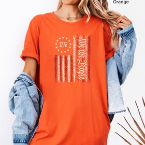 May include: Orange t-shirt featuring a distressed American flag design. The flag includes the text "1776" in a circle of stars and the words "We the People" vertically. The shirt has short sleeves and a relaxed fit.