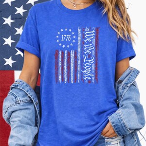 May include: Blue t-shirt featuring a distressed American flag design. The flag includes the text "1776" within a circle of stars and the words "We the People" in a vertical column. The shirt is worn with a denim jacket.
