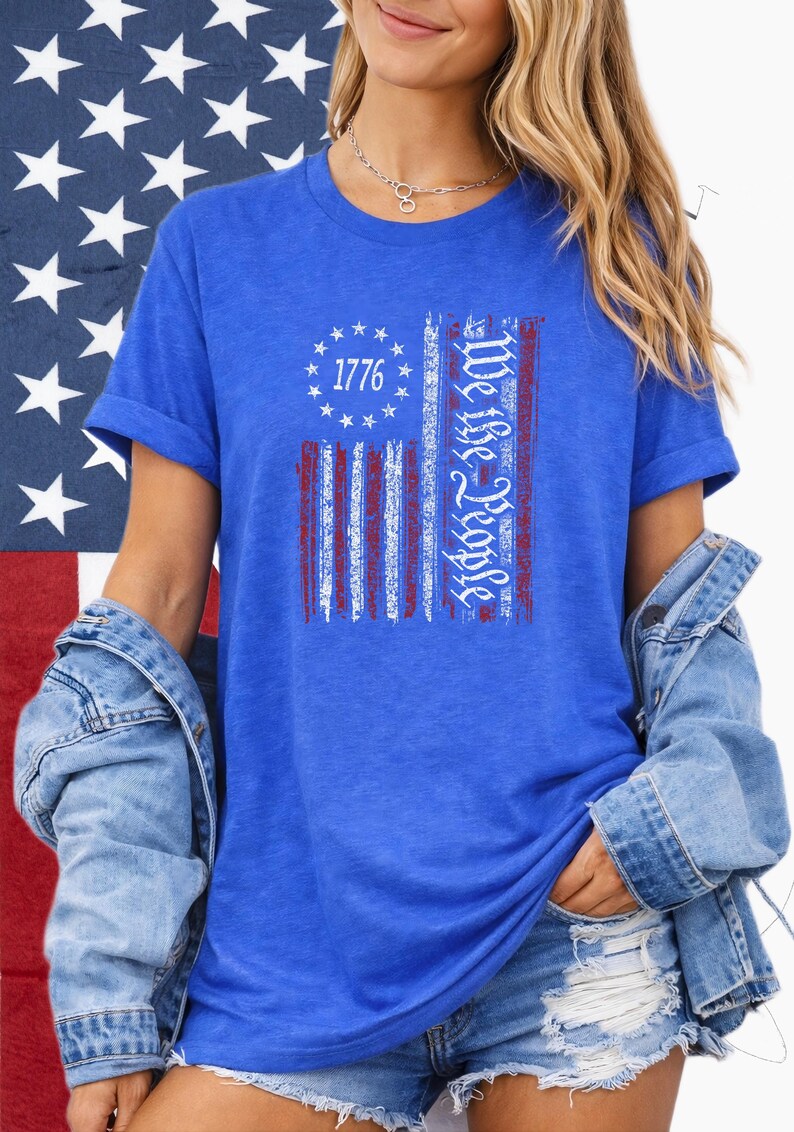 May include: Blue t-shirt featuring a distressed American flag design. The flag includes the text "1776" within a circle of stars and the words "We the People" in a vertical column. The shirt is worn with a denim jacket.