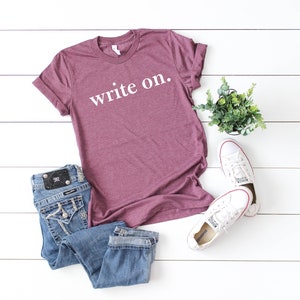 May include: A maroon t-shirt with the text "write on." in white lettering. The shirt is laid flat on a white surface with a pair of blue jeans, white sneakers, and a potted plant.
