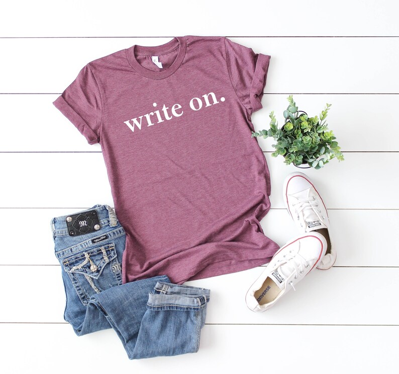 May include: A maroon t-shirt with the text "write on." in white lettering. The shirt is laid flat on a white surface with a pair of blue jeans, white sneakers, and a potted plant.
