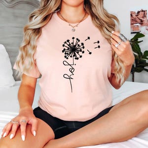 K&ouml;nnte beinhalten: Ein pinkfarbenes T-Shirt mit einem schwarzen L&ouml;wenzahnmuster und dem Wort "Freude" in einer Schreibschrift.