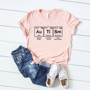 May include: A pink t-shirt with a graphic of the periodic table elements Au, Ti, and Sm. The graphic includes the atomic number, atomic weight, and element name for each element. The text below each element describes its properties. The shirt is laid out on a white wooden surface with a pair of white sneakers and blue jeans.