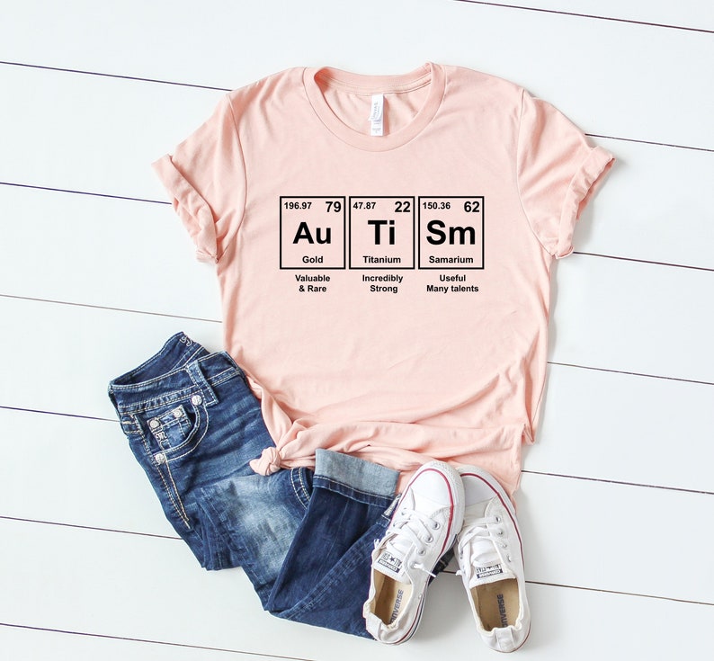 May include: A pink t-shirt with a graphic of the periodic table elements Au, Ti, and Sm. The graphic includes the atomic number, atomic weight, and element name for each element. The text below each element describes its properties. The shirt is laid out on a white wooden surface with a pair of white sneakers and blue jeans.