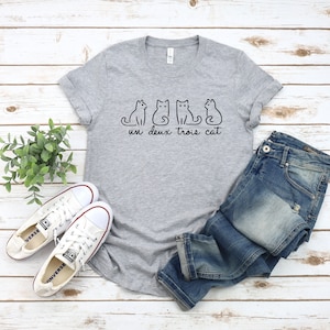 May include: Gray t-shirt with a graphic of four cats and the text "un deux trois cat". The shirt is paired with blue jeans and white sneakers.