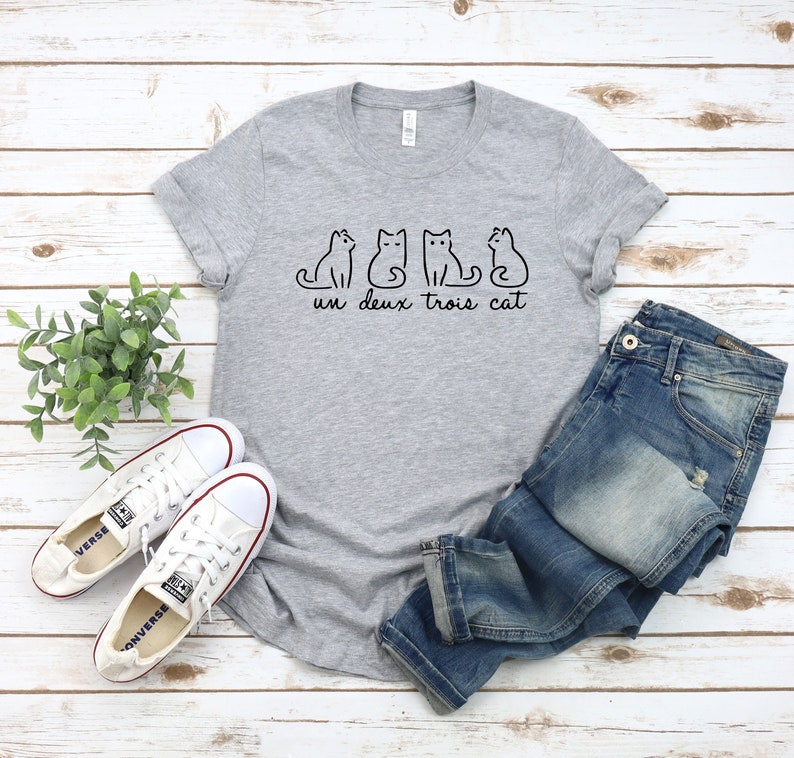 May include: Gray t-shirt with a graphic of four cats and the text "un deux trois cat". The shirt is paired with blue jeans and white sneakers.