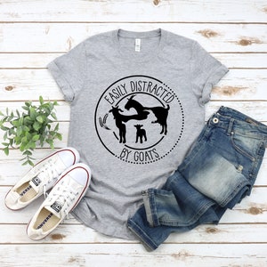 May include: A grey t-shirt with a black circle graphic that says "Easily Distracted by Goats". The graphic features three goats, two adults and a kid, in a circle with wheat stalks.