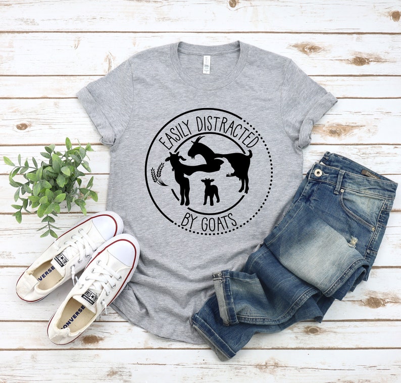 May include: A grey t-shirt with a black circle graphic that says "Easily Distracted by Goats". The graphic features three goats, two adults and a kid, in a circle with wheat stalks.