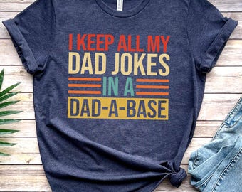 May include: Heather navy t-shirt with the text "I KEEP ALL MY DAD JOKES IN A DAD-A-BASE" in red, beige, orange, and teal. The shirt is folded and has a knot at the bottom. A pair of blue jeans and a brown shoe are visible.