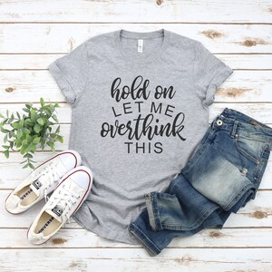 Pode incluir: Uma camiseta cinza com o texto "hold on let me overthink this" em letras pretas. A camiseta &eacute; combinada com jeans azul e t&ecirc;nis brancos.