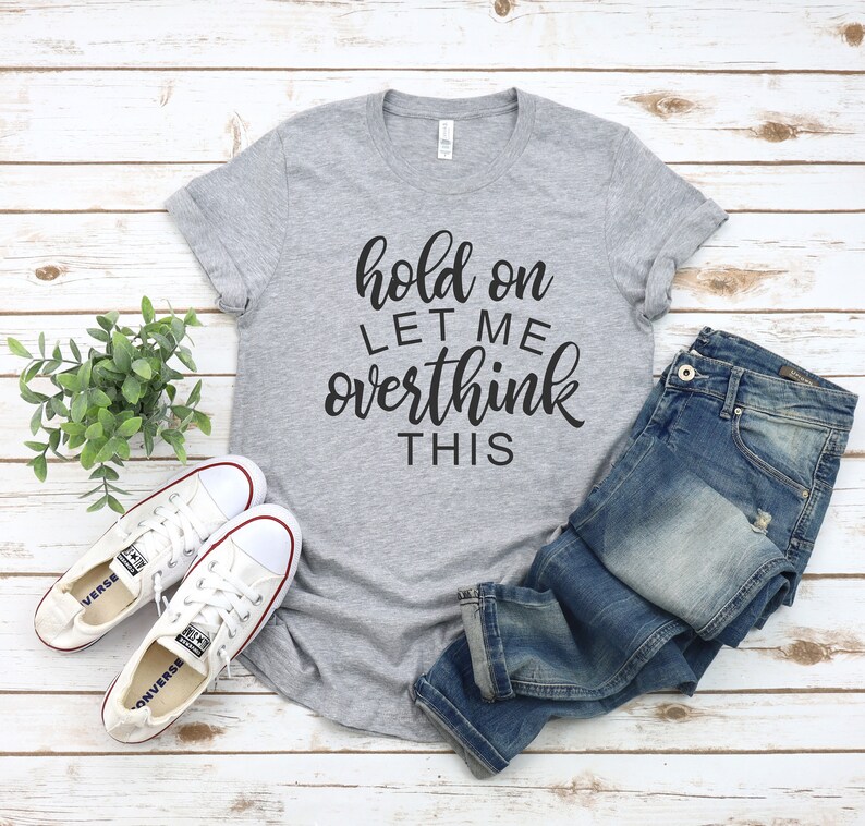 Pode incluir: Uma camiseta cinza com o texto "hold on let me overthink this" em letras pretas. A camiseta &eacute; combinada com jeans azul e t&ecirc;nis brancos.