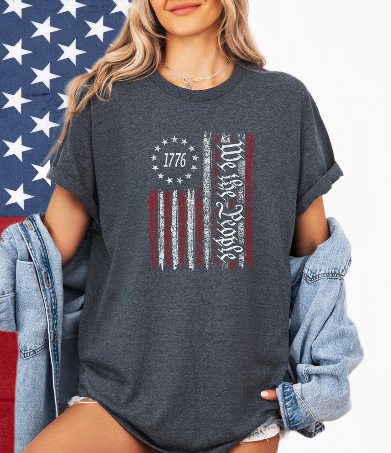 May include: Dark gray t-shirt featuring a distressed American flag design. The flag includes the text "1776" and "We the People" in white. The shirt is a crew neck and has short sleeves.