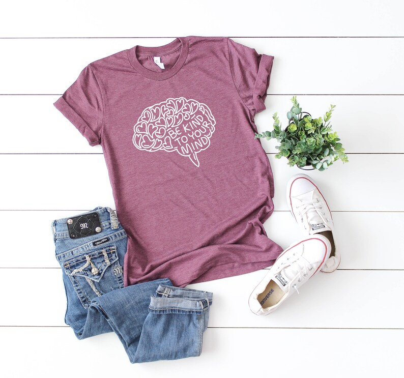 May include: A maroon t-shirt with a white outline of a brain that says "Be Kind To Your Mind". The shirt is laying on a white wooden surface with a pair of white sneakers, blue jeans, and a small potted plant.