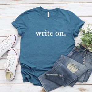 May include: A teal blue t-shirt with the text "write on." printed in white. The t-shirt is laid flat on a white wooden surface with a pair of white sneakers and blue jeans.