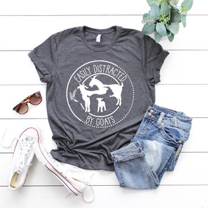 May include: A dark grey t-shirt with a white circle design that says "Easily Distracted by Goats". The design features three goats, two adults and a kid, in a playful pose. The shirt is laid out on a white wooden surface with a pair of jeans, sunglasses, and white trainers.