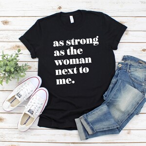 May include: Black t-shirt with white text that reads "as strong as the woman next to me."
