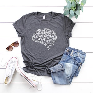 May include: A dark gray t-shirt with a white outline of a brain with the text "Be Kind To Your Mind" inside. The shirt is laid out on a white wooden surface with a pair of sunglasses, white sneakers, and blue jeans.