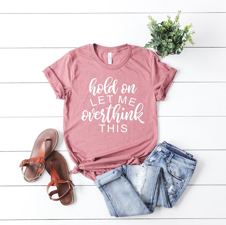 May include: A pink t-shirt with the text 'hold on let me overthink this' in white lettering. The t-shirt is laid out on a white wooden surface with a pair of brown sandals and blue jeans.