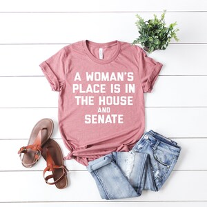 May include: A pink t-shirt with the text "A woman's place is in the house and senate" in white lettering.