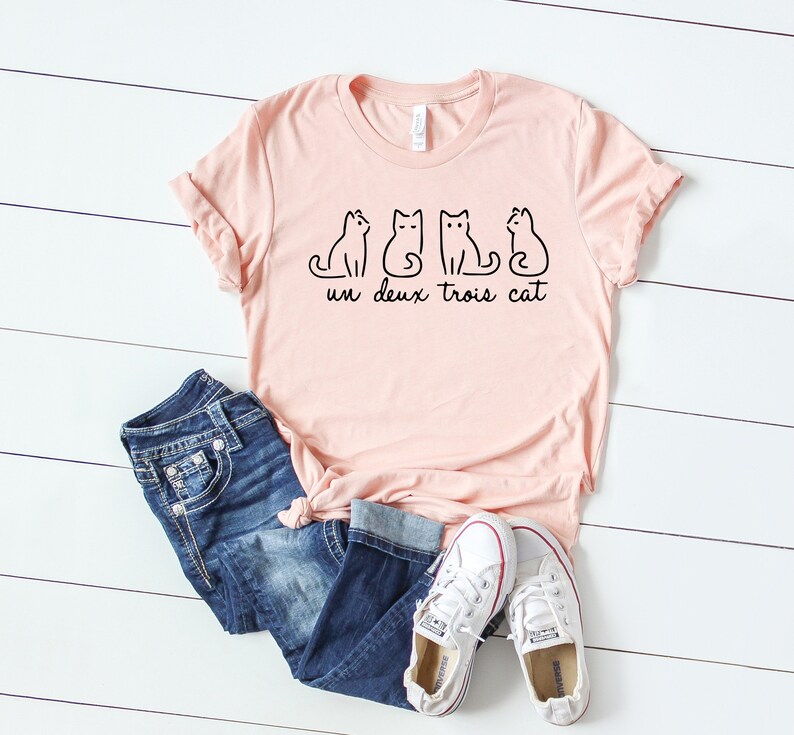 May include: A pink t-shirt with a graphic of three cats and the text "un deux trois cat". The shirt is paired with blue jeans and white sneakers.