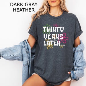 May include: Dark heather grey t-shirt with white text that reads "Thirty Years Later..." surrounded by colorful cartoon-style flowers.