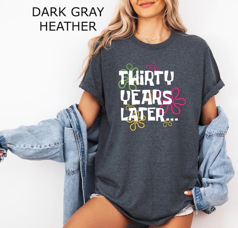 May include: Dark heather grey t-shirt with white text that reads "Thirty Years Later..." surrounded by colorful cartoon-style flowers.