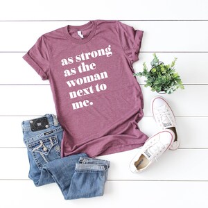 May include: A maroon t-shirt with the text "as strong as the woman next to me." in white lettering. The shirt is laid out on a white wooden surface with a pair of white trainers, a potted plant, and a pair of blue jeans.