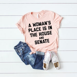 May include: A pink t-shirt with black text that reads "A woman's place is in the house and senate".
