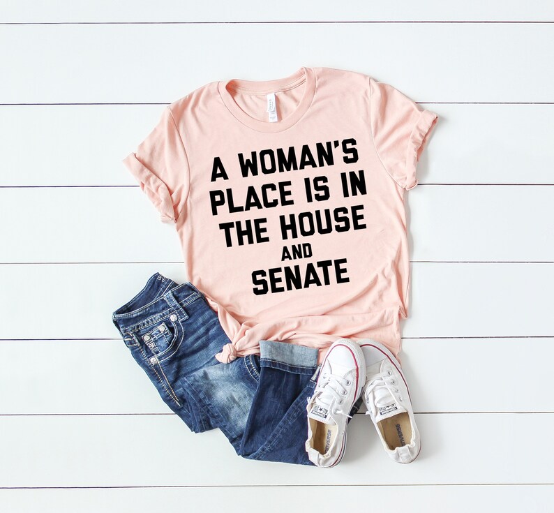 May include: A pink t-shirt with black text that reads "A woman's place is in the house and senate".