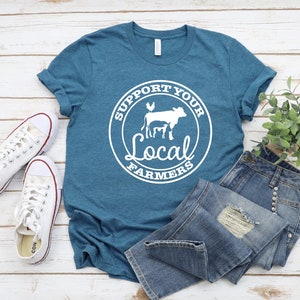May include: A teal blue t-shirt with a white circle graphic that says "Support Your Local Farmers" with a cow, calf, and chicken inside the circle. The t-shirt is laying on a white wooden surface with a pair of white sneakers and blue jeans.