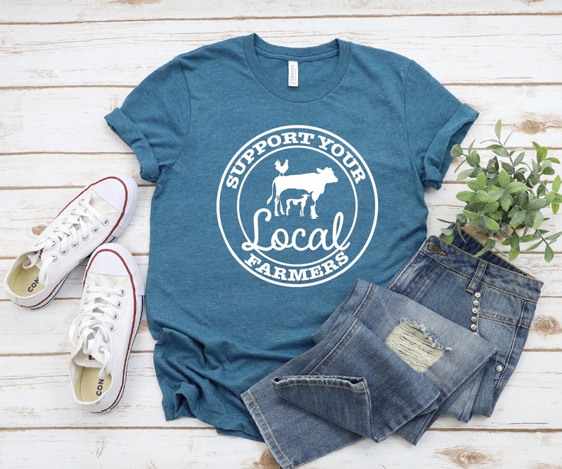 May include: A teal blue t-shirt with a white circle graphic that says "Support Your Local Farmers" with a cow, calf, and chicken inside the circle. The t-shirt is laying on a white wooden surface with a pair of white sneakers and blue jeans.
