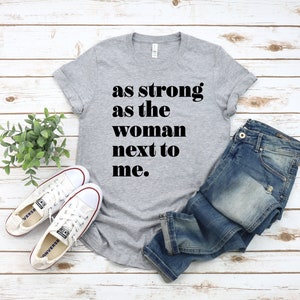 May include: Grey t-shirt with the text "as strong as the woman next to me."