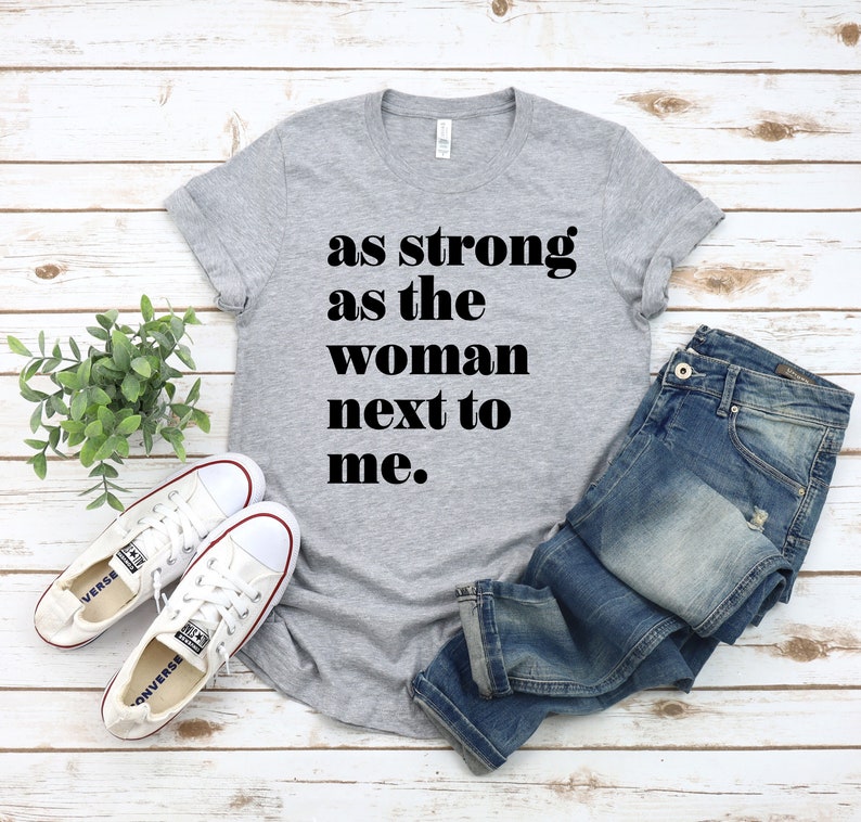 May include: Grey t-shirt with the text "as strong as the woman next to me."