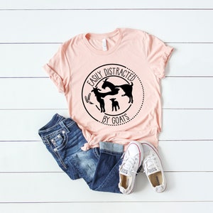 May include: A pink t-shirt featuring a black circle graphic that reads "Easily Distracted by Goats". The graphic depicts a silhouette of three goats. The shirt is styled with blue jeans and white trainers.