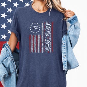 May include: Navy blue t-shirt featuring a distressed American flag design. The flag includes the text "1776" and "We the People" in white script. The shirt has short sleeves and a relaxed fit.