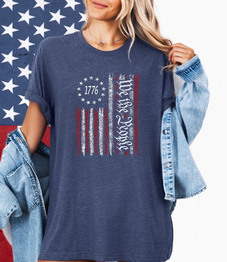 May include: Navy blue t-shirt featuring a distressed American flag design. The flag includes the text "1776" and "We the People" in white script. The shirt has short sleeves and a relaxed fit.