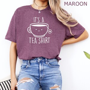 May include: A heather maroon t-shirt with a white graphic of a teacup with a smiling face and the text "It's a Tea Shirt".