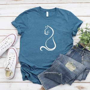 May include: A teal blue t-shirt with a white outline of a cat sitting on the front. The t-shirt is laying on a white wooden surface with a pair of white sneakers and a pair of blue jeans.
