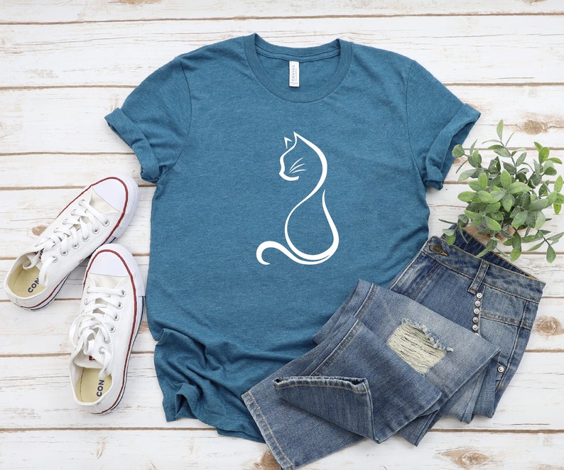 May include: A teal blue t-shirt with a white outline of a cat sitting on the front. The t-shirt is laying on a white wooden surface with a pair of white sneakers and a pair of blue jeans.