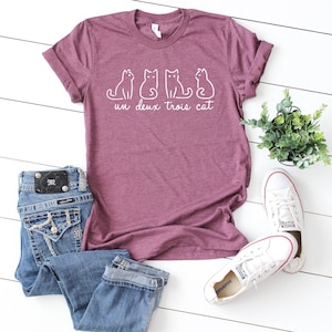 May include: A maroon t-shirt with a white graphic of three cats and the text "un deux trois cat".