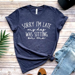 Op de afbeelding: Een donkerblauw T-shirt met witte tekst die "Sorry, ik ben te laat, mijn hond zat op me" zegt.