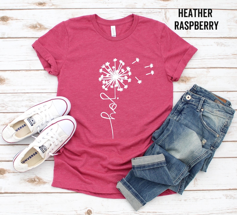 K&ouml;nnte beinhalten: Ein T-Shirt in der Farbe Heidelbeer-Himbeere mit einem wei&szlig;en L&ouml;wenzahnmuster und dem Wort "joy" darunter. Das Shirt wird mit einer blauen Jeans und wei&szlig;en Turnschuhen kombiniert.