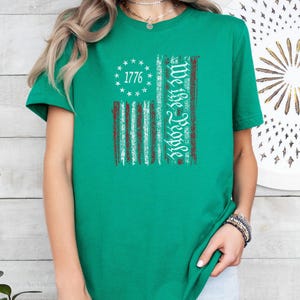 May include: A green t-shirt featuring a distressed American flag design. The flag includes the text "1776" surrounded by stars and the words "We the People" in a vertical column. The shirt is a solid green color.