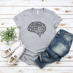 May include: Gray t-shirt with a black outline of a brain with the text "Be Kind To Your Mind" inside. The shirt is laid out on a white wood surface with a pair of white sneakers and blue jeans.